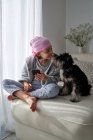 Сверху очаровательный больной мальчик в розовой бандане и пижаме ласкает питомца, сидя дома на кровати, используя мобильный телефон — стоковое фото