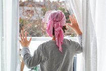 Vista posterior del niño anónimo con cáncer usando pañuelo rosa y poniendo las manos en la ventana en la habitación - foto de stock