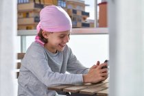 Criança alegre com doença oncológica curtindo passatempo com celular no terraço — Fotografia de Stock