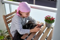 Fröhliches kleines krebskrankes Kind genießt Zeitvertreib mit Handy auf der Terrasse, während es einen kleinen Hund hält — Stockfoto
