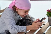 Cheerful little child with cancer disease enjoying pastime with cellphone on terrace while holding a small dog — Stock Photo