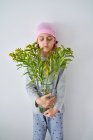 Focused little boy with cancer diagnosis wearing pink bandana with closed eyes while holding vase with flowers and standing at wall — Stock Photo