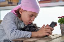 Cheerful little child with cancer disease enjoying pastime with cellphone on terrace while holding a small dog — Stock Photo