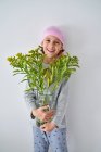 Cheerful little boy with cancer diagnosis wearing pink bandana and looking at camera while holding vase with flowers and standing at wall — Stock Photo