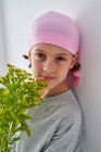 Fröhlicher kleiner Junge mit Krebsdiagnose trägt rosa Kopftuch und blickt in die Kamera, während er eine Vase mit Blumen in der Hand hält und an der Wand steht — Stockfoto