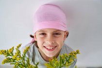 Menino alegre com diagnóstico de câncer vestindo bandana rosa e olhando para a câmera enquanto segurando vaso com flores e de pé na parede — Fotografia de Stock