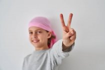 Petit enfant courageux avec diagnostic de cancer en regardant la caméra faire un geste de victoire avec les doigts sur fond gris — Photo de stock