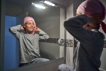 Вид сзади больного ребенка, надевающего розовую бандану перед зеркалом в ванной комнате — стоковое фото