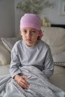 Grave bambino carino in bandana rosa guardando la fotocamera e combattendo il cancro a casa seduto in un divano — Foto stock