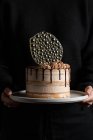 Personne méconnaissable tenant un gâteau festif avec remplissage et glaçage au chocolat et décoration en isomalt d'argent sur table avec fond noir — Photo de stock