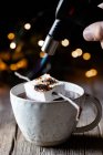 Анонимный человек с кухонным горелкой, обжаривающий зефир на палочке над элегантной чашкой горячего шоколада на деревянном столе с блестками на темном фоне — стоковое фото