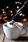 Anonymous person with kitchen burner roasting marshmallow placed on stick over elegant cup of hot chocolate on wooden table with sparkles in dark background — Stock Photo
