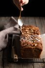 Crop housewife with spoon in hand pouring white sugar icing over homemade banana bread with nuts placed on metal grid on wooden table — Stock Photo