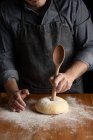 Crop male baker in black apron using big wooden spoon for making hole in dough while forming artisan round bread loaf at wooden table — Stock Photo