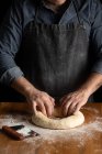 Crop male baker in black apron making hole in dough while forming artisan round bread loaf at wooden table — Stock Photo