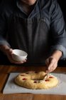 Crop chef en tablier noir graissant pain rond non cuit avec jaune d'oeuf surmonté de cerise tout en se tenant debout à la table en bois — Photo de stock