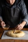 Crop chef masculino en delantal negro engrasar pan redondo sin cocer con yema de huevo mientras está de pie en la mesa de madera - foto de stock