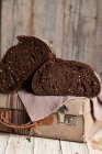 Pan de centeno oscuro saludable apetitoso con granos cortados por la mitad colocados en la maleta de tela retro en la mesa de madera en mal estado - foto de stock