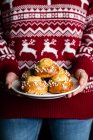 Кукурудзяна самиця в червоному різдвяному светрі і джинсах тримає тарілку з домашнім тістечком і демонструє смачні свіжі булочки з білими зморшками — стокове фото