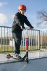 Aktiver Teenie-Junge mit Schutzhelm und Tretroller steht auf Rampe im Skatepark, während er sich auf Tricks an sonnigen Frühlingstagen vorbereitet — Stockfoto