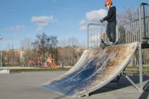 From below of active teen boy in protective helmet with kick scooter standing on ramp in skate park while preparing for performing trick in sunny spring day — Stock Photo