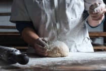 Personne méconnaissable pétrissant la pâte avec de la farine sur la table tout en travaillant dans la boulangerie — Photo de stock
