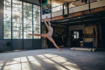 Вид сбоку на юную стройную балерину в спортивном костюме, прыгающую с парашютом, практикующую танцевальные движения в светлой просторной студии с большим окном — стоковое фото