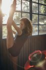 Vista lateral da jovem dançarina com os olhos fechados em bodysuit preto de pé perto da janela e alongamento da perna enquanto se exercita em estúdio — Fotografia de Stock