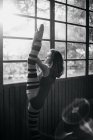 Side view of young female dancer in black bodysuit standing near window and stretching leg while exercising in studio — Stock Photo
