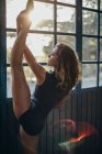 Side view of young female dancer looking away in black bodysuit standing near window and stretching leg while exercising in studio — Stock Photo