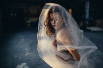 Молодая талантливая танцовщица с закрытыми глазами в купальнике танцует с прозрачной вуалью, тренируясь в одиночестве в светлой студии — стоковое фото