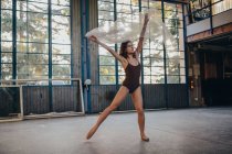 Танцовщица в черном купальнике и пуантах танцует с прозрачным светлым тюлем во время репетиции в студии — стоковое фото