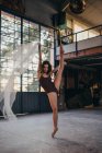 Corpo pieno di sottile graziosa ballerina guardando la fotocamera in body nero e scarpe da punta che eseguono danza con tulle leggero trasparente durante le prove in studio — Foto stock