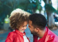 Side view of adorable cheerful ethnic girl embracing happy father wearing similar leather jacket while resting together in park in sunny day — Stock Photo