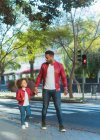 Счастливый молодой этнический человек с маленькой дочерью, одетый в аналогичный наряд, держась за руки во время прогулки по улице города в солнечный день — стоковое фото
