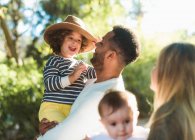 Joyeux jeune famille multiraciale avec de petits enfants profiter du temps ensemble tout en se reposant dans un parc verdoyant dans une journée d'été ensoleillée — Photo de stock