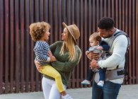 Cheerful stylish young multiracial couple with infant son and little daughter in casual wear walking together along wooden fence on city street — Stock Photo