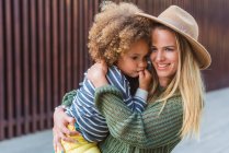 Side view of cheerful young female in trendy sweater and hat embracing cute little daughter while standing on street near modern fence — Stock Photo