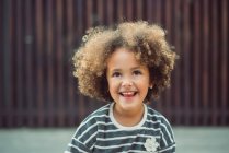 Adorable little girl with curly hair wearing casual striped shirt smiling while standing against blurred wall on street — Stock Photo