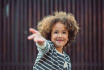 Adorabile bambina con i capelli ricci che indossa camicia a righe casual sorridente mentre in piedi contro il muro sfocato sulla strada — Foto stock