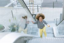 From above of cute ethnic curly haired girl in trendy outfit standing on stair of escalator and eating apple in city — Stock Photo
