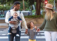 Happy young multiracial parents with little daughter eating apple and cute infant boy crossing road on zebra while walking in city in summer day — Stock Photo