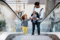 Сверху положительный молодой этнический мужчина несет младенца и держит за руку веселую маленькую дочь, стоя на эскалаторе в городе — стоковое фото