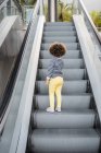 Vista trasera de chica de pelo rizado étnico lindo irreconocible en traje de moda de pie en la escalera de escaleras mecánicas en la ciudad - foto de stock