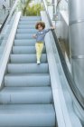 From below of cute ethnic curly haired girl in trendy outfit standing on stair of escalator and eating apple in city — Stock Photo