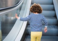 Back view of unrecognizable cute ethnic curly haired girl in trendy outfit standing on stair of escalator in city — Stock Photo