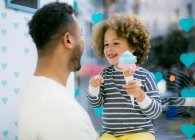 Encantadora chica étnica rizada sosteniendo cono dulce de helado jugando con el padre amoroso negro en la cafetería - foto de stock