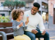 Homem negro feliz olhando para a filha encantadora desfrutando de cone de sorvete doce enquanto descansa no banco na praça da cidade — Fotografia de Stock