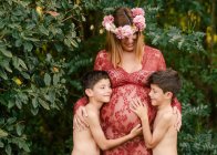 Счастливая мать в красивом красном платье и цветочный венок улыбаясь и обнимая близнецов сыновей обнимая живот, стоя возле зеленых деревьев в саду в летний день — стоковое фото