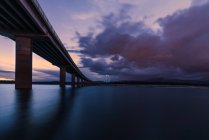 Lange Brücke überquert ruhigen Fluss gegen bewölkten Himmel mit Blitz am Abend in der Landschaft — Stockfoto